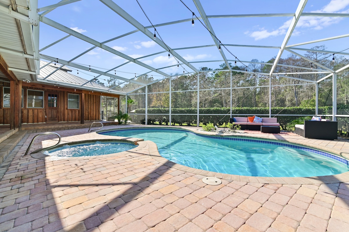 An enclosed outdoor area features a sparkling swimming pool with a built-in hot tub, surrounded by a stone patio. Comfortable seating arrangements are visible along one side, and greenery can be seen beyond the screening, contributing to the serene ambiance.