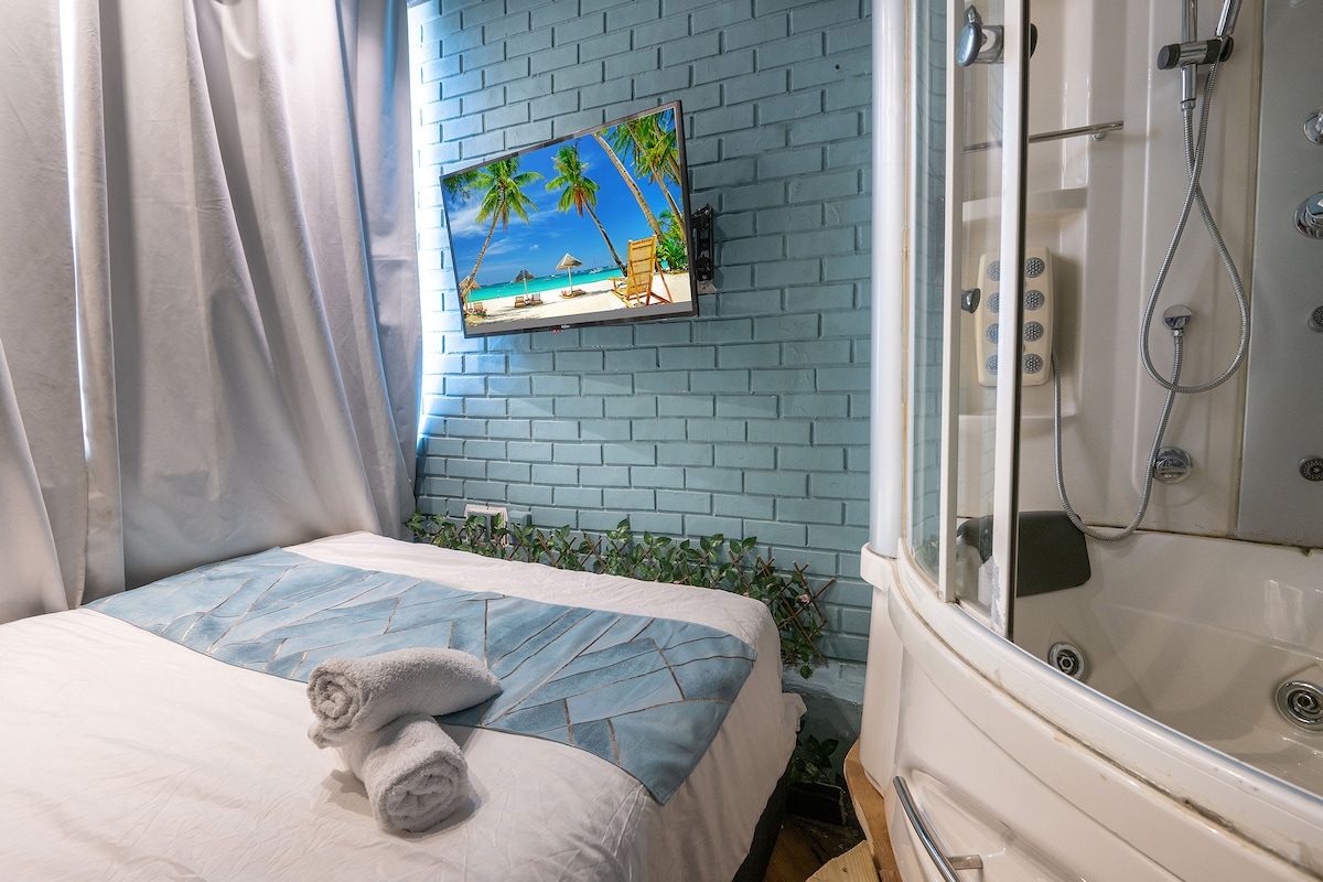 A modern room features a spacious bed draped with a blue blanket, accompanied by neatly rolled towels. A large window allows natural light to enter, while a flat-screen television displays a beach scene. The en-suite bathroom includes an American-style shower and a jacuzzi.