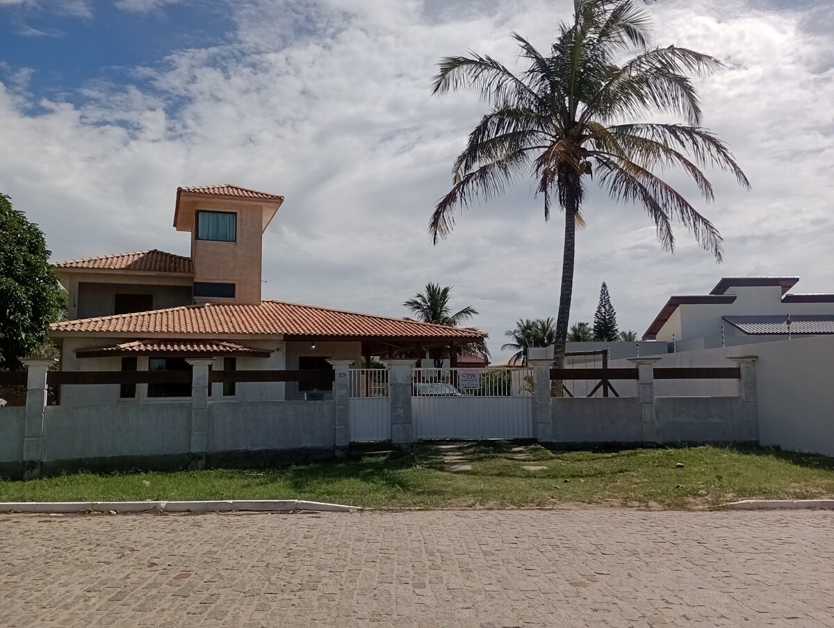 A spacious house is featured, showcasing a rooftop terrace and a tiled roof. Palm trees and other greenery surround the exterior. Fencing encloses the property, with a gated entrance visible from the street. The sky is partly cloudy, enhancing the serene setting.