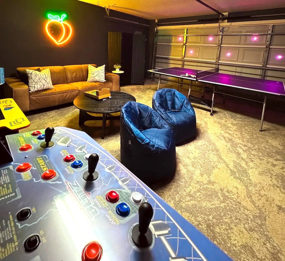 A vibrant game room features a classic arcade game with colorful controls and buttons. Cozy bean bag chairs are arranged around a circular coffee table, while a ping pong table sits nearby. Neon accent lights enhance the playful atmosphere in the space.