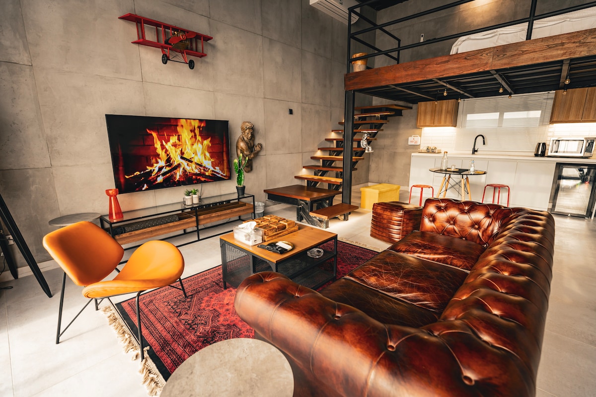 A loft-style living area is highlighted by a leather couch and a vintage rug. A flat-screen television displays a fireplace image, while a staircase leads to the upper floor bedroom. The industrial decor is complemented by wooden accents and stylish furnishings.