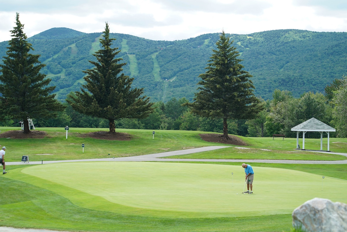 A golfer practices putting on a well-maintained green, surrounded by expansive lawns and tall trees. Distant mountains provide a scenic backdrop, with ski trails visible on the slopes. A gazebo is situated nearby, enhancing the serene atmosphere of the golf course.