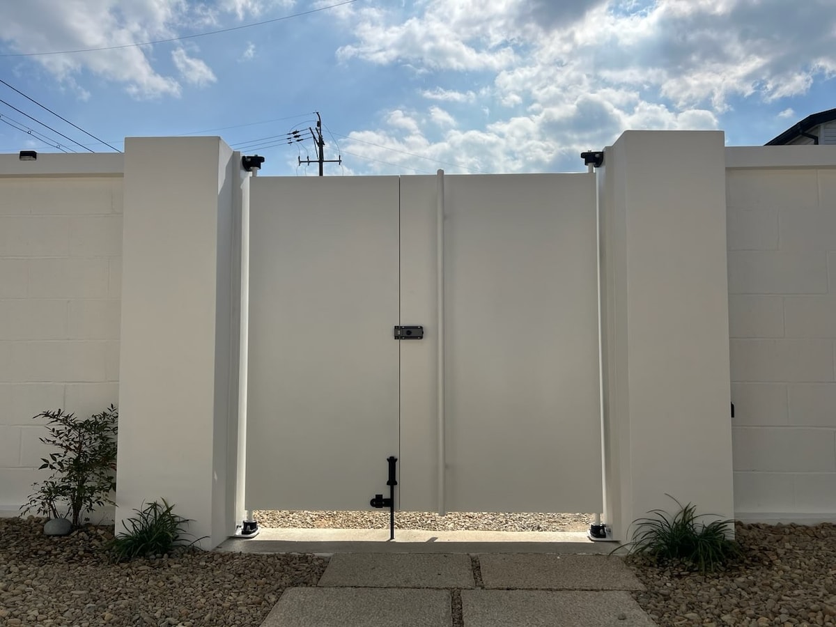 A modern, white gate is centered within a low, white wall, leading to a tidy courtyard. The gate features a simple handle and a secure latch. Around the gate, small plants are situated on either side, with a gravel pathway visible beneath a partially cloudy sky.