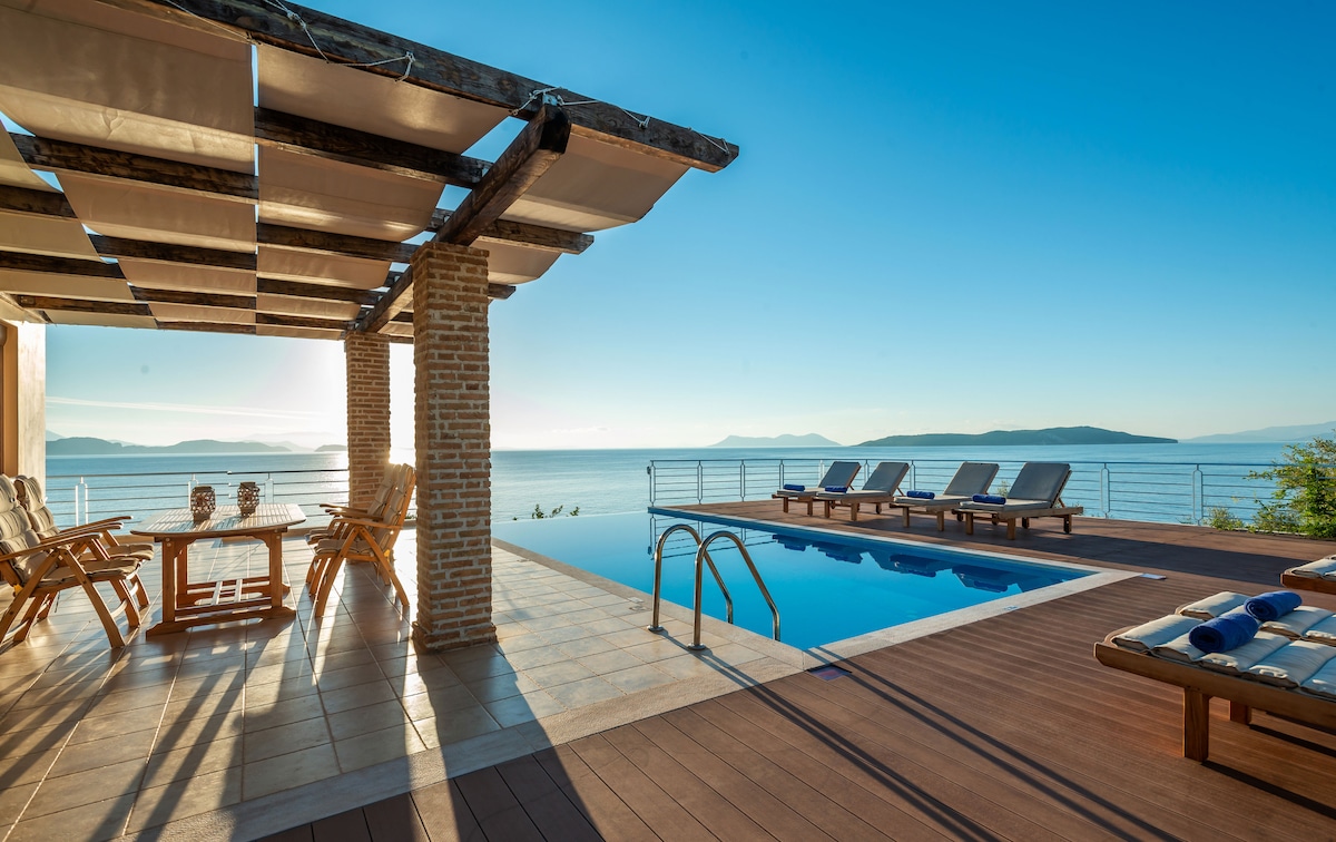 An inviting outdoor space is showcased with a swimming pool and lounge chairs positioned to enjoy panoramic sea views. A shaded seating area with wooden furniture complements the sun-drenched deck, with sunlight casting gentle shadows on the tiles. The horizon features tranquil water meeting the sky.