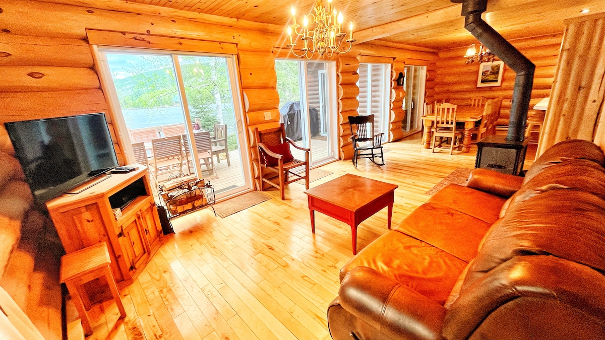 An inviting living area features natural wood walls and a cozy seating arrangement. A television is positioned nearby, with a coffee table centrally located. Large sliding doors open onto a deck, offering views of the lake. A rustic chandelier completes the ambiance.