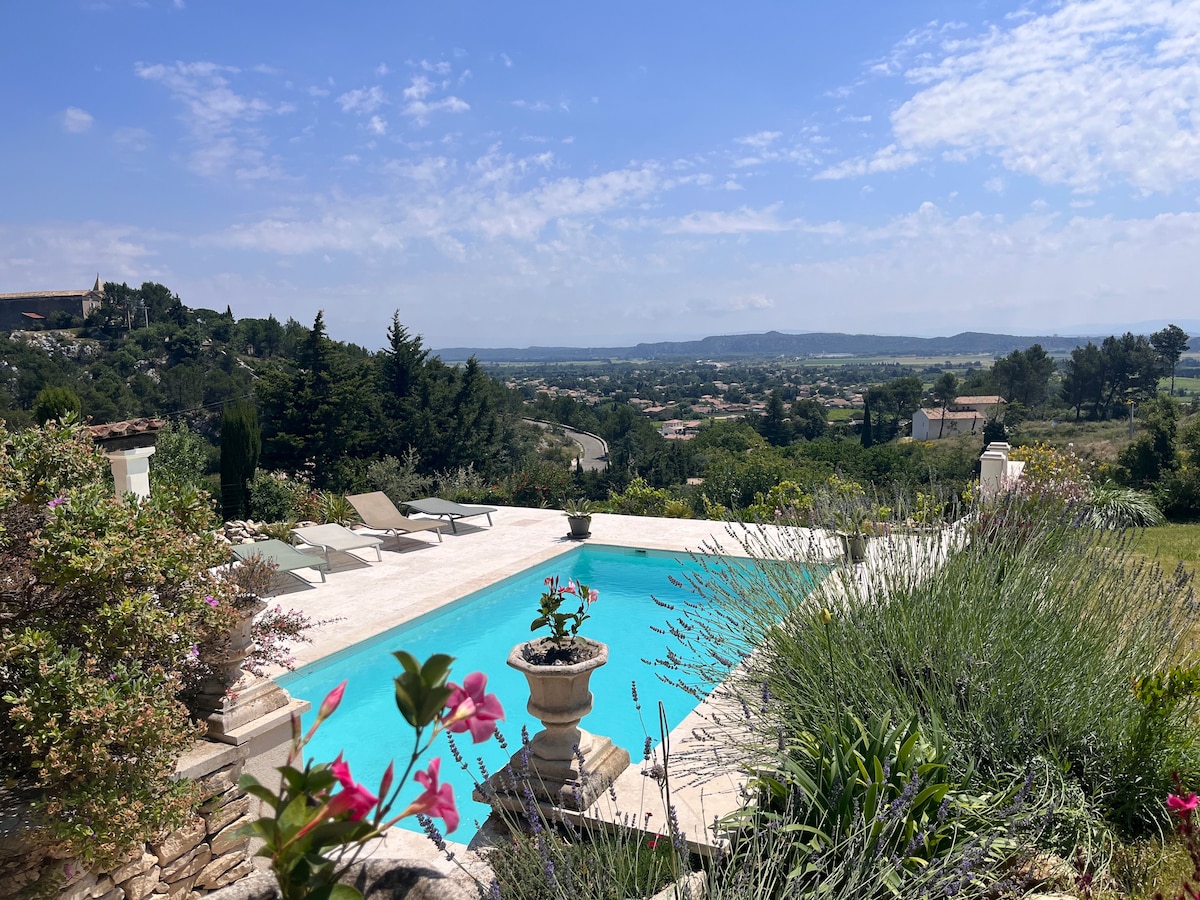 A serene view of the saltwater pool surrounded by lush greenery and blooming flowers. Loungewear chairs are positioned near the pool's edge, offering a perfect spot to relax. Rolling hills stretch into the distance under a clear blue sky with gentle clouds.