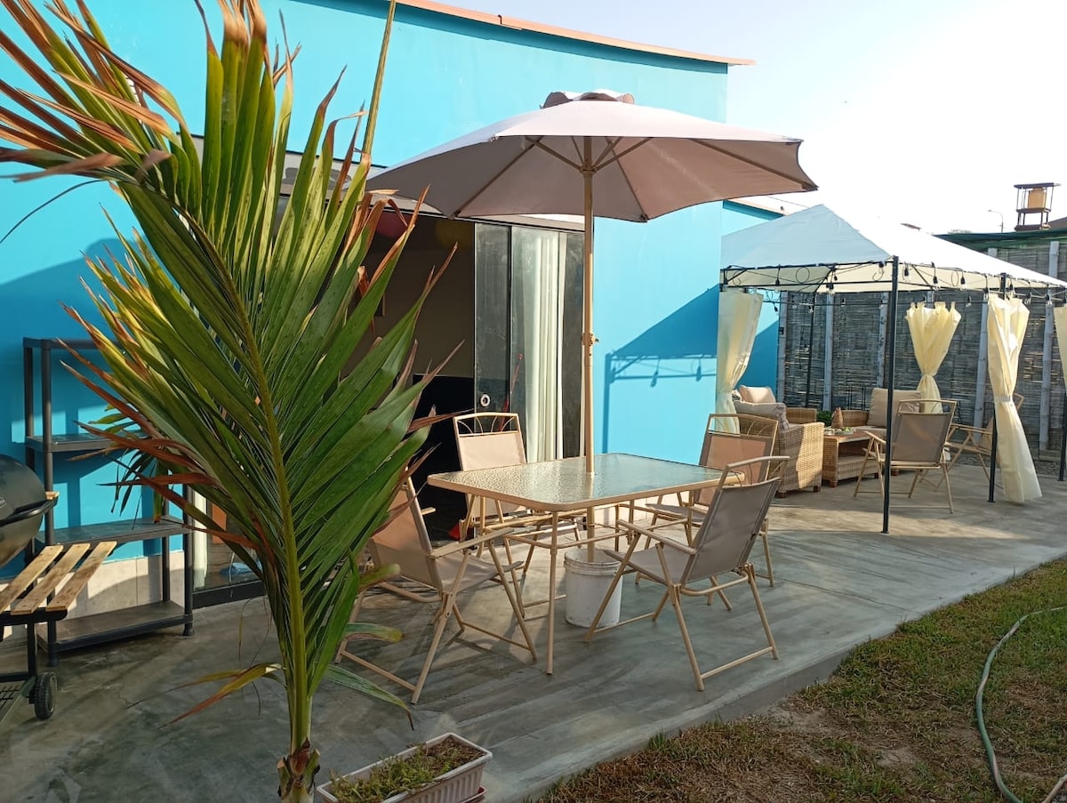 An outdoor patio area features a large table surrounded by several light-colored chairs. A large umbrella provides shade, and a gazebo is positioned to the side. A palm plant adds greenery to the space, contributing to a relaxed outdoor atmosphere.