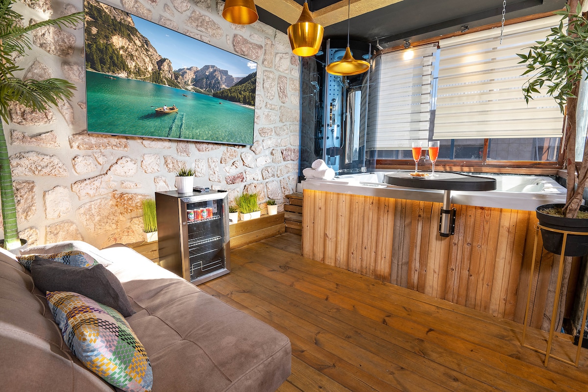 An inviting spa area features a wooden hot tub next to a soft sofa with decorative pillows. A large wall-mounted screen displays a scenic landscape. Natural light filters through partially closed blinds, creating a warm ambiance. Indoor plants add a touch of nature.