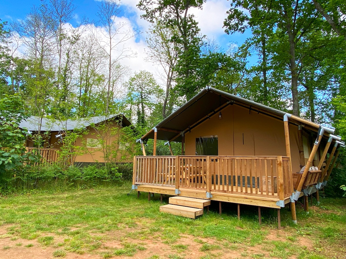 Two lodge tents are nestled among greenery, featuring wooden decks and steps for easy access. The structures are made of high-quality materials, showcasing a modern yet rustic design. Sunlight filters through the trees, creating a serene outdoor atmosphere.