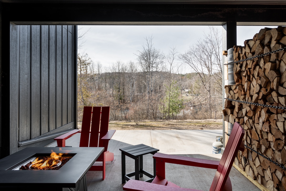 A cozy outdoor seating area features two red adirondack chairs facing a modern fire pit. A small black side table complements the seating arrangement. Stacked firewood is seen nearby, with a serene view of nature visible through the open space.