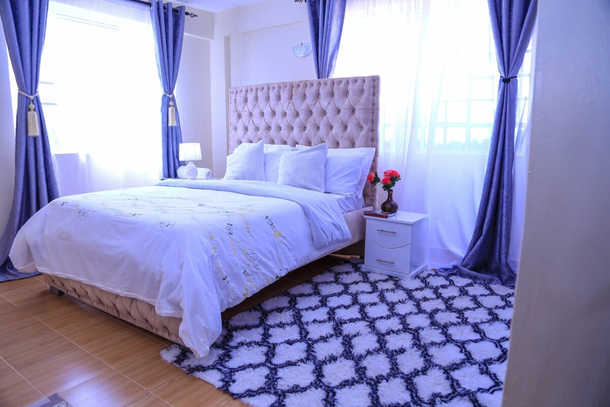 A spacious sleeping area features a tufted headboard with a light-colored bedspread adorned with delicate embroidery. Curtains in a soft blue frame the window, allowing natural light to illuminate the room. A small nightstand holds a vase with fresh flowers beside the bed, enhancing the serene atmosphere.