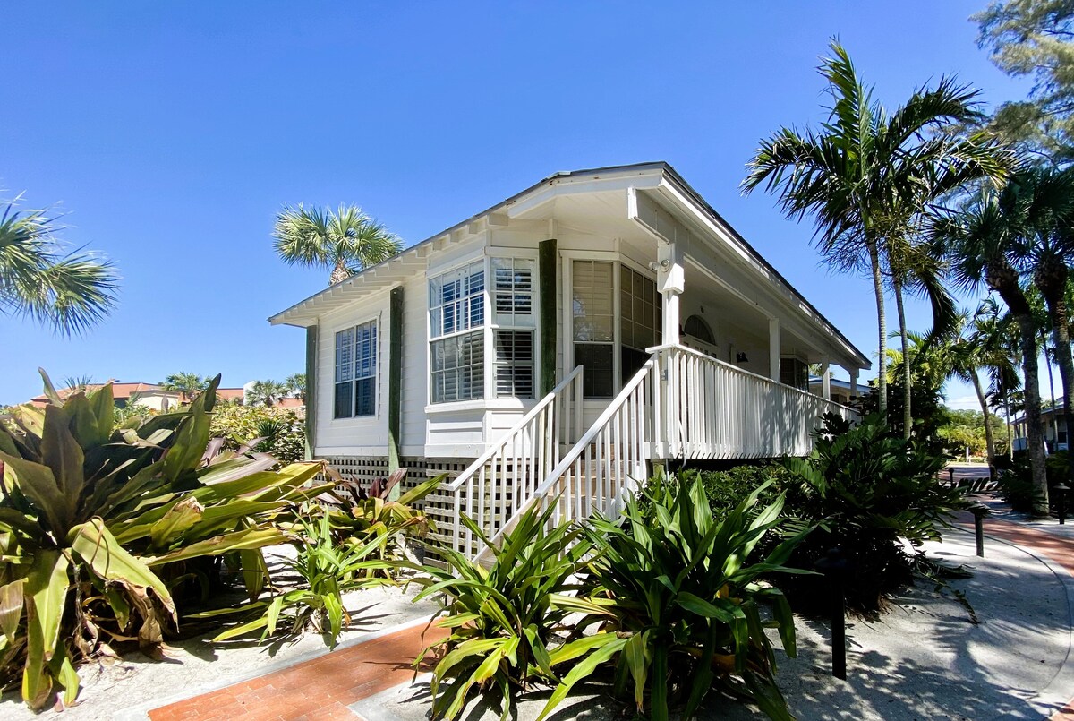 A charming coastal cottage is surrounded by lush green foliage and palm trees, showcasing white siding and a welcoming front porch. Steps lead to the entrance, framed by vibrant shrubs, against a clear blue sky.