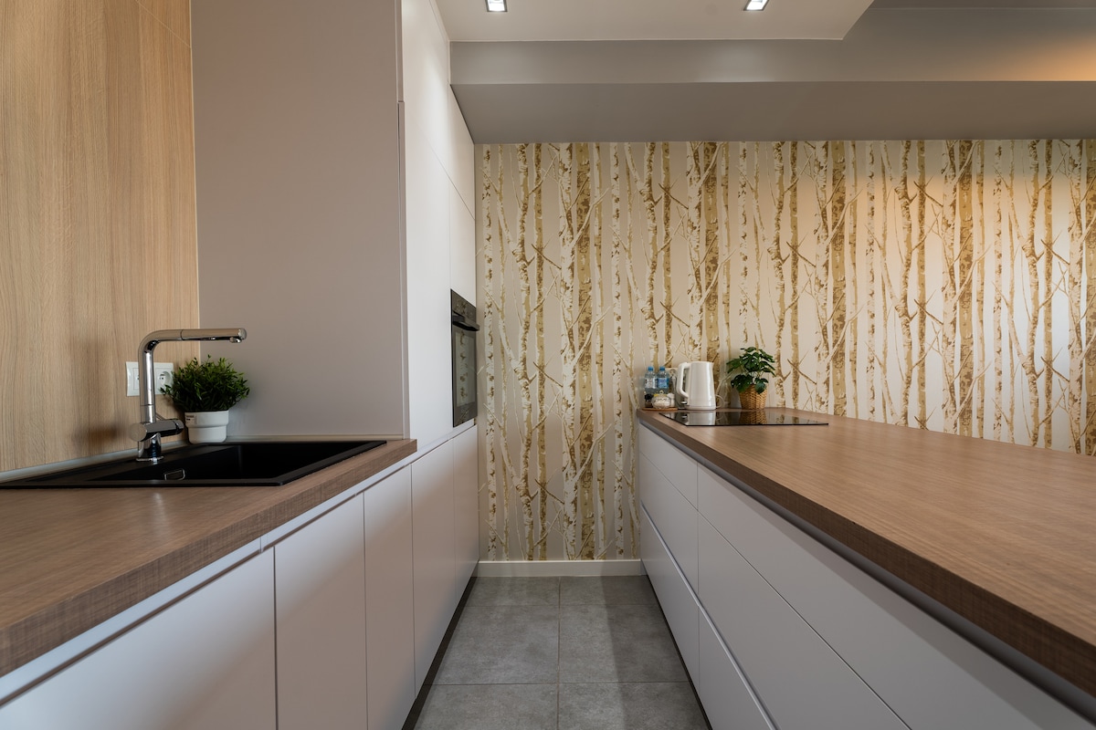 A modern kitchen is presented with sleek cabinetry and a spacious counter. Soft wood tones create a warm atmosphere against the patterned wallpaper. A black sink is integrated into the cabinetry, complemented by various kitchen essentials placed on the counter.