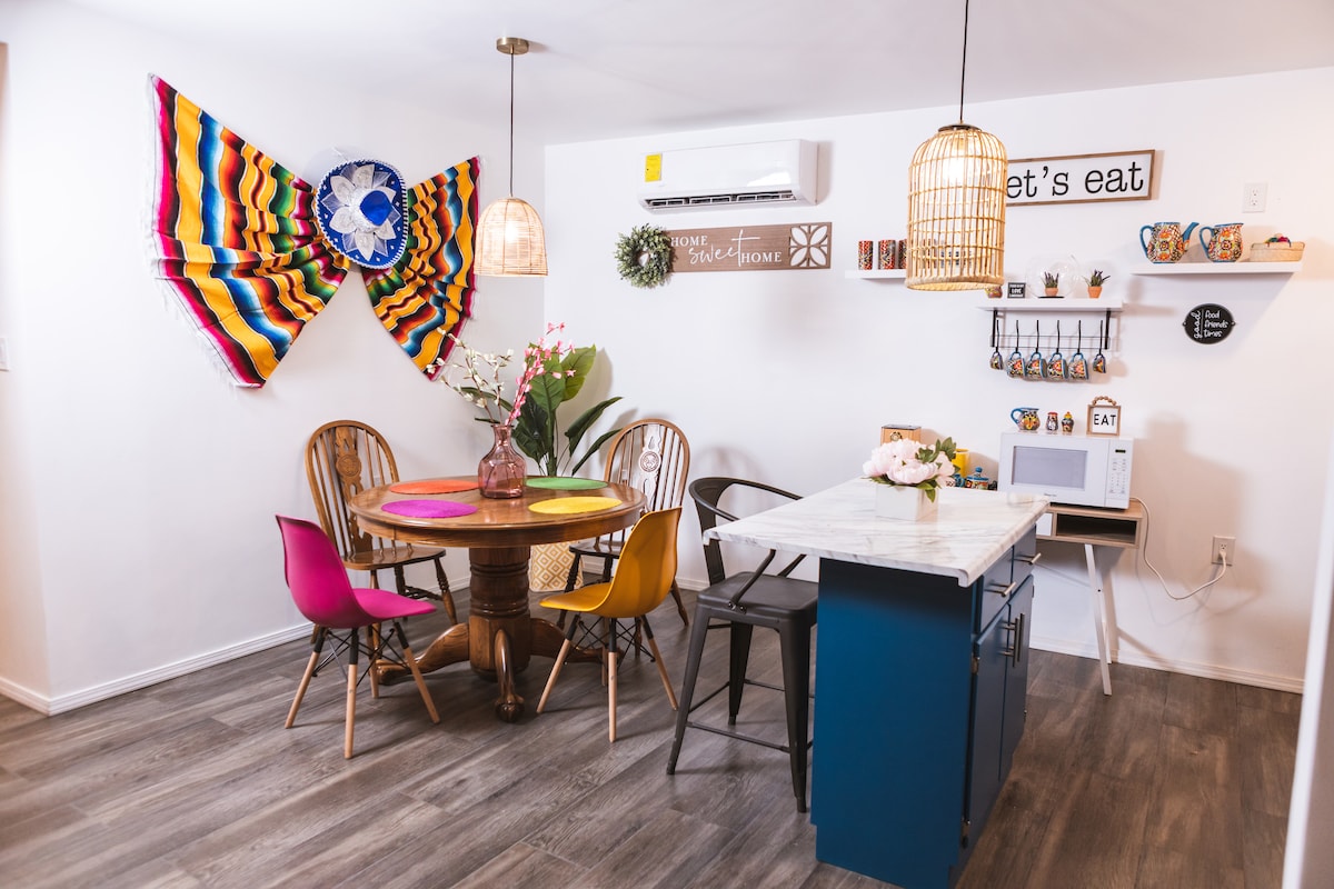 A dining area features a round wooden table surrounded by colorful chairs in pink, yellow, and natural wood tones. Bright textiles are displayed on the wall, and potted plants add a touch of greenery. An air conditioning unit and microwave are visible for guest convenience.