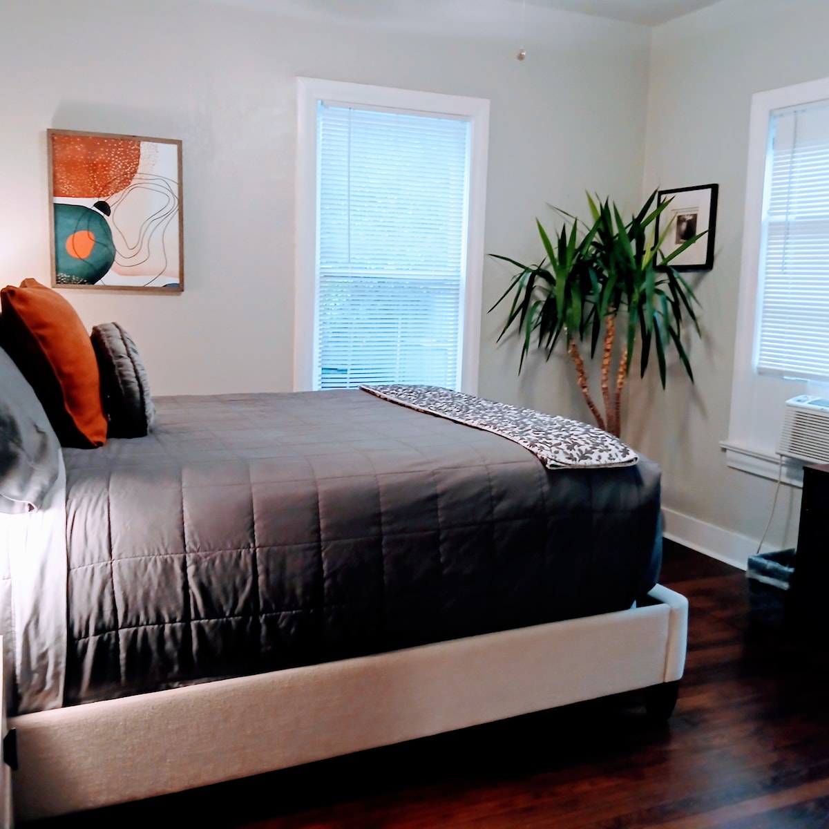 A well-lit bedroom features a spacious bed with neutral bedding and accent pillows. A leafy plant is placed near the window, allowing natural light to fill the room. Artwork decorates the wall, adding a touch of creativity to the calming space.