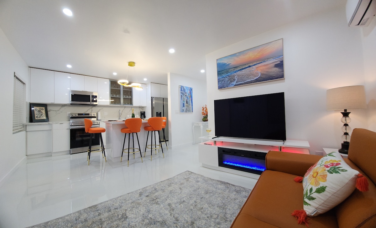 A modern living space is showcased with a sleek kitchen and dining area featuring orange bar stools. A comfortable brown sofa is positioned beside a stylish entertainment unit with ambient lighting. Artwork depicting a beach landscape adorns the wall, complementing the bright, open layout.