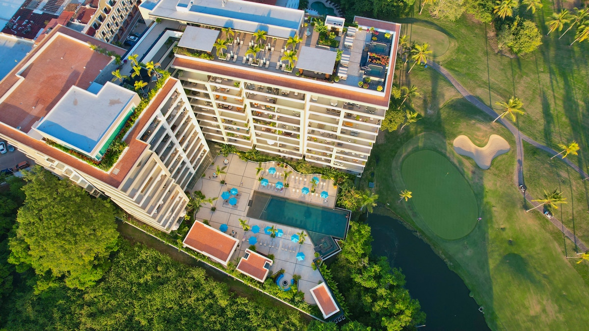 An aerial perspective captures a modern condominium with a spacious swimming pool, surrounded by sun loungers. The lush greenery of the golf course extends beside the building, featuring palm trees and a manicured putting green. Rooftop terraces are visible, enhancing the building's contemporary design.