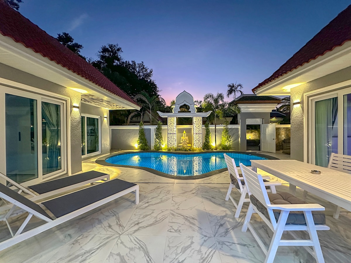 An outdoor area is highlighted with a private pool reflecting warm light from outdoor illumination. Lounge chairs are arranged beside the pool, and a dining table is positioned nearby. Lush greenery surrounds the space, creating a tranquil environment at dusk.
