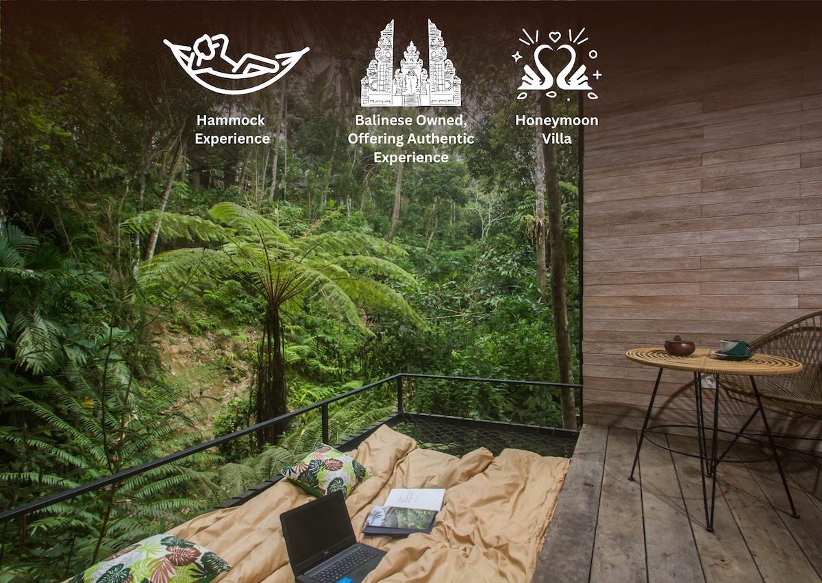 A cozy terrace features a spacious daybed adorned with a light brown blanket. Lush green foliage surrounds the area, creating a sense of tranquility. A small table is placed nearby with a laptop and two wicker chairs, inviting relaxation and connection with nature.