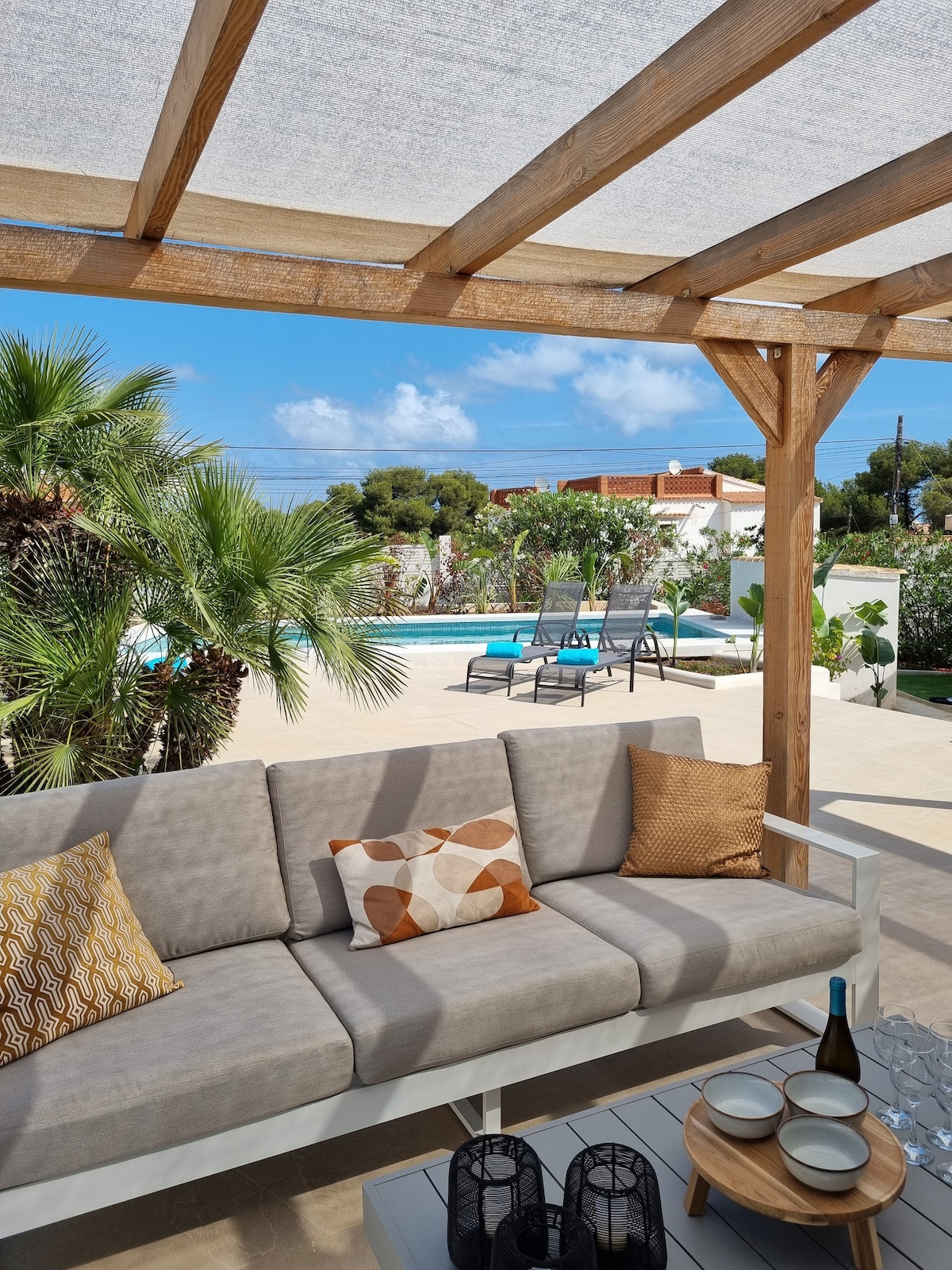 An outdoor lounge area features a light-colored sofa adorned with decorative cushions. A table is set with a bottle, glasses, and lanterns. The backdrop showcases a private swimming pool surrounded by lush greenery and sun loungers, under a shaded pergola.