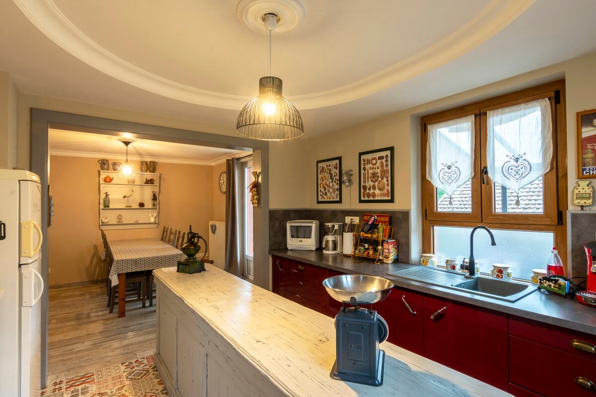 A modern kitchen features sleek red cabinetry and a large central island with a decorative scale. Natural light fills the room through two windows adorned with lace curtains, while a dining area with a checkered tablecloth is visible in the background.