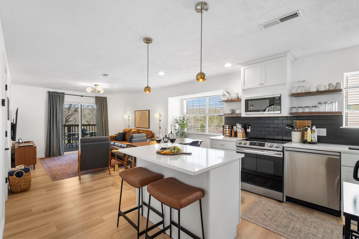 A modern kitchen is showcased, featuring a large island with seating for two. Stainless steel appliances and elegant cabinetry provide functionality. The open space leads to a cozy living area, illuminated by natural light, with a comfortable seating arrangement and large windows offering views of the outdoors.