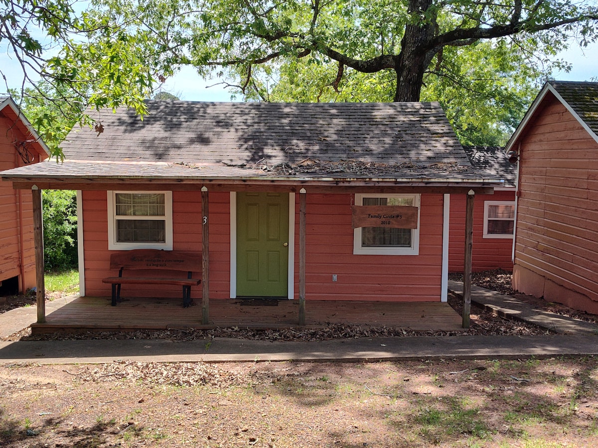 A cabin with a welcoming entrance features a green door set against pink siding. A wooden bench is placed on the porch, under the shade of a large tree, providing a comfortable outdoor sitting area. The structure is surrounded by greenery.