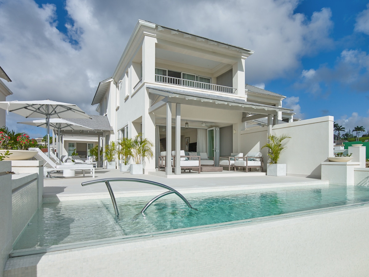 A two-story villa is displayed, featuring a modern architectural design with light-colored walls. The exterior includes a private pool with a sleek water feature. A sheltered patio is visible alongside lounge seating and an umbrella, with lush greenery enhancing the surroundings.