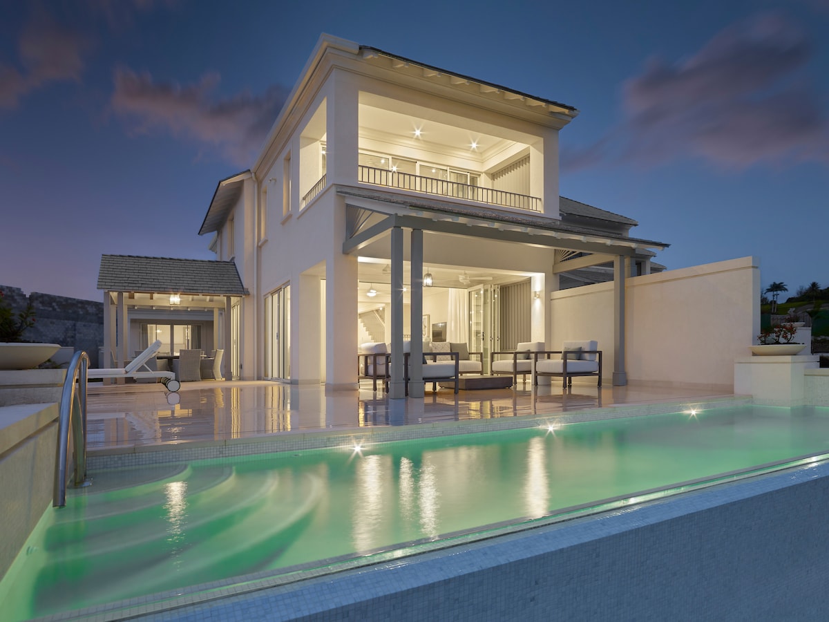 The two-story villa showcases a modern architectural design with expansive glass doors, enhancing the connection between indoor and outdoor spaces. A private pool glows softly in the twilight, reflecting the serene ambiance of the surrounding area.