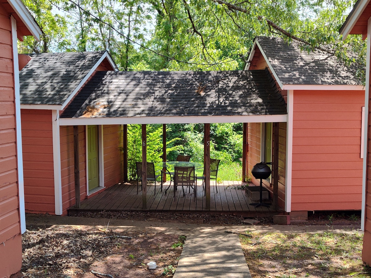 A cabin exterior is showcased, featuring a covered porch with a wooden deck. A patio table and chairs are positioned under the protective roof, while a grill is placed nearby. Lush greenery surrounds the structure, creating a natural backdrop.