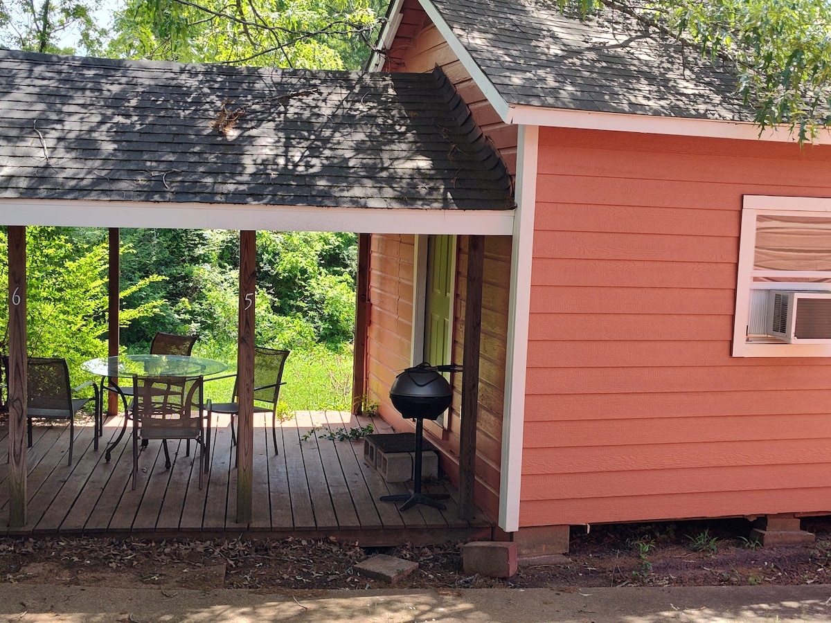 A charming cabin exterior is shown, featuring peach-colored walls and a shaded porch. A dining table with four chairs is positioned nearby, accompanied by a grill on the side. Lush greenery can be seen in the background, enhancing the serene environment.