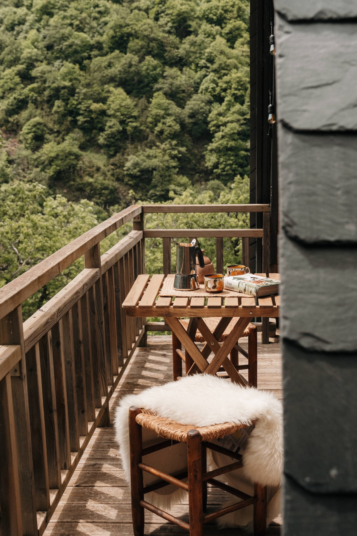 A wooden terrace is visible, surrounded by lush greenery. A small, rustic table is set with a coffee pot and a variety of breakfast items. Two inviting chairs, one adorned with a soft, white fur throw, contribute to the comfortable outdoor dining atmosphere.