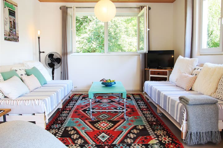 Tauche ein in warmes Licht, umgeben von vielen Fenstern zum Garten. Genieße die Gemütlichkeit der großen Sofas mit kuscheligen Kissen und dem handgewebten bosnischen Cilim – stilvoll und einladend.
