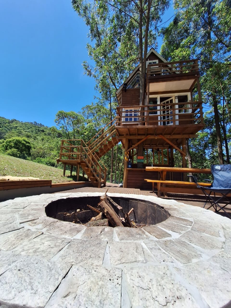 A charming treehouse is visible, set among tall trees under a bright blue sky. A circular stone fire pit is situated in the foreground, with logs placed for a fire. Wooden stairs lead up to the treehouse, inviting exploration.