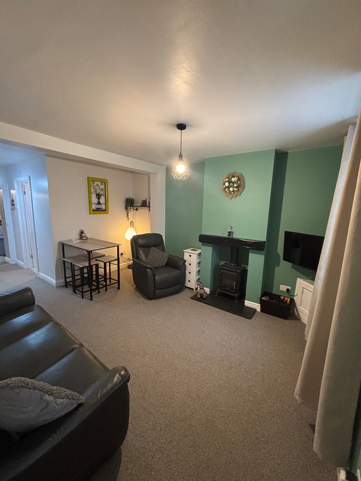 A living area features a dark leather sofa and a cozy armchair positioned near the log burner. A small dining/work table sits in the corner. The walls are painted in a calming green, illuminated by a pendant light, while a TV is mounted on one wall.