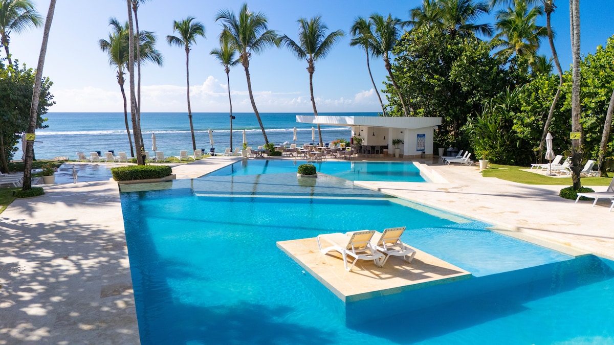 An expansive swimming pool is surrounded by palm trees, offering a view of the ocean. Sun loungers are positioned around the pool area, creating a serene environment. The calm waters reflect the clear sky, inviting relaxation and enjoyment of the outdoor space.