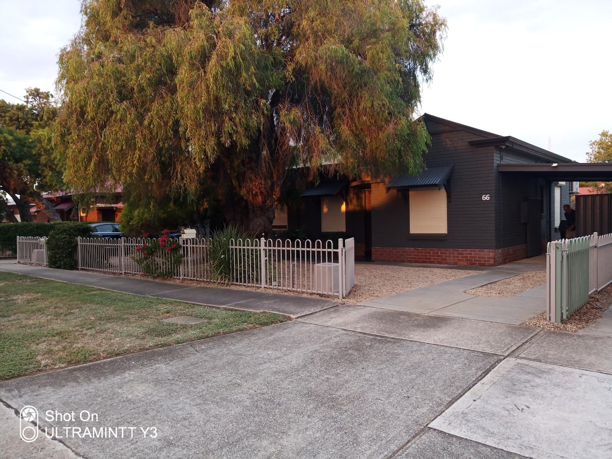 The exterior of a single-story home is shown, featuring a neatly landscaped yard with a gravel pathway. A large tree provides shade in front, while a fenced area borders the property. Warm light glows from the windows, creating a welcoming impression.
