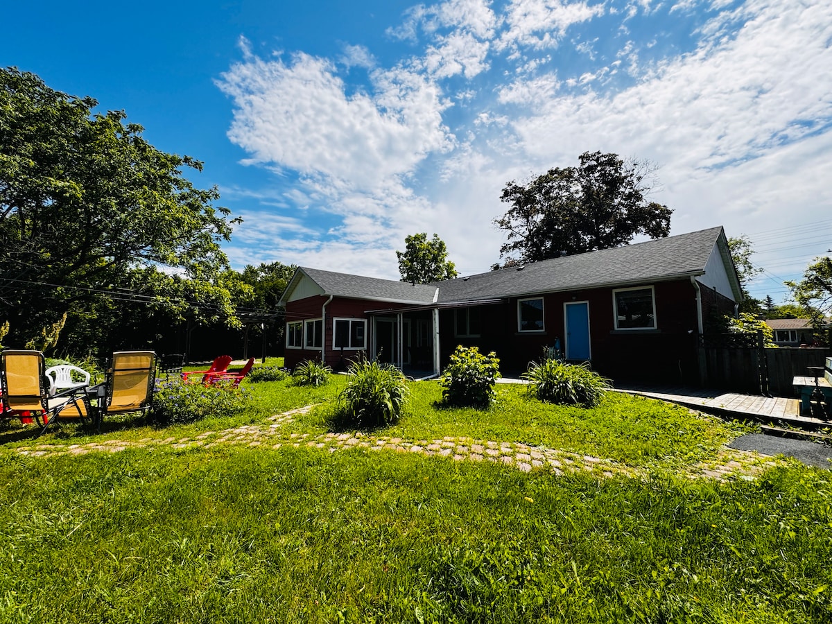 The home's exterior is shown against a clear blue sky, featuring a landscaped yard with grass and various plants. A walking path leads to the entrance, while patio furniture, including chairs, is arranged on one side for outdoor relaxation.