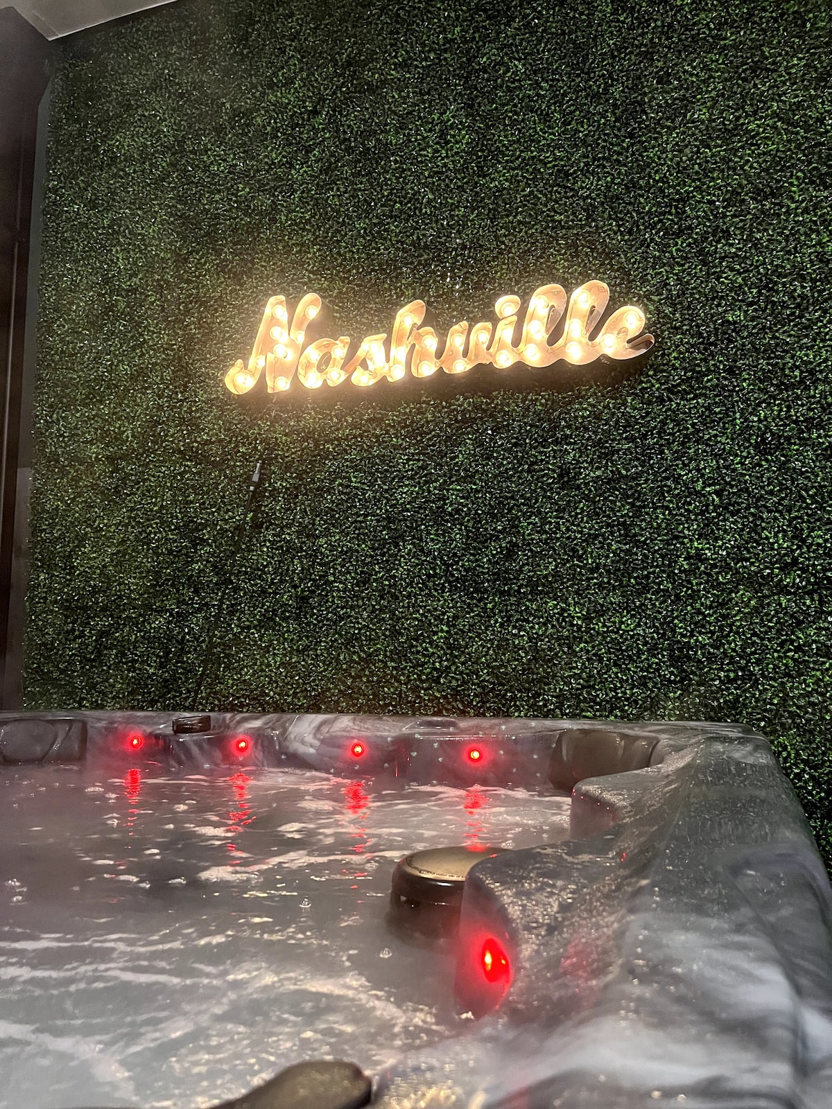 A hot tub with illuminated jets invites relaxation, surrounded by a green, textured wall. Above the hot tub, a glowing sign reading 'Nashville' adds a touch of local charm, enhancing the space’s warm ambiance.