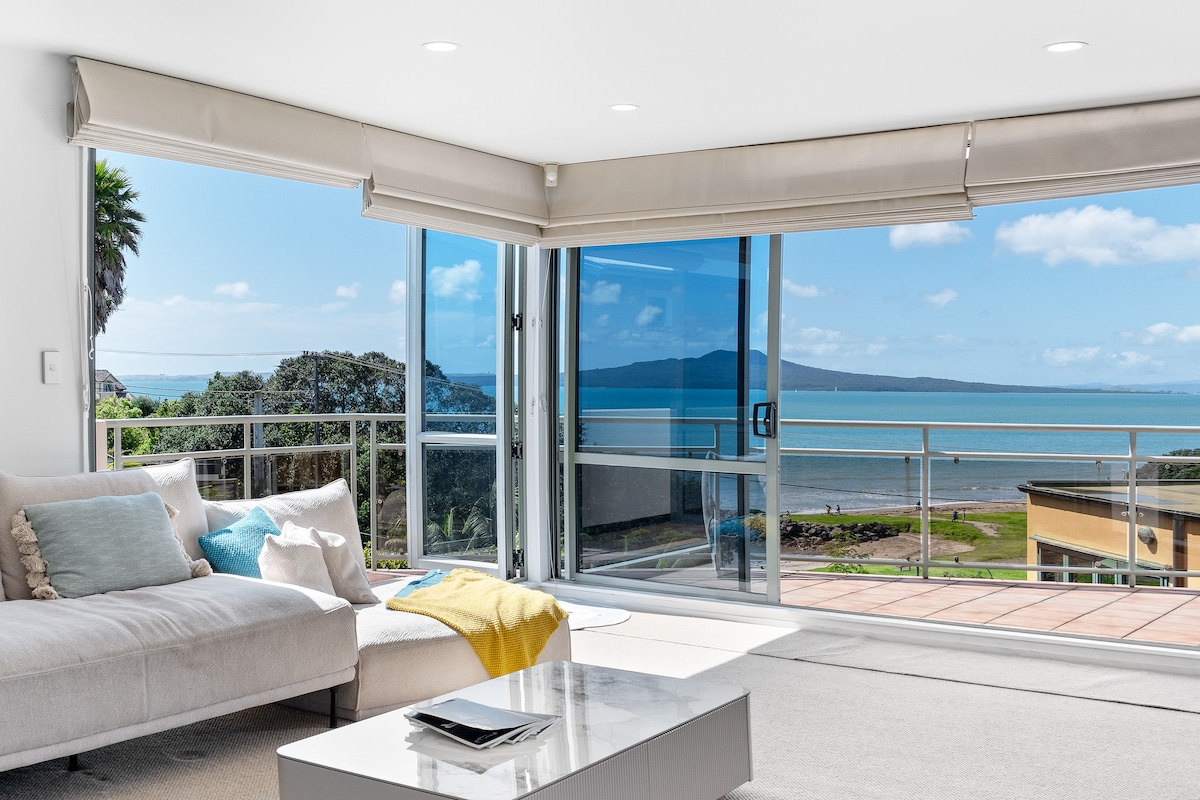 A bright living area is showcased, featuring large sliding glass doors that open to reveal expansive sea views. A light-colored sofa adorned with decorative cushions faces a sleek coffee table. Natural light pours in, enhancing the spacious feel of the room.