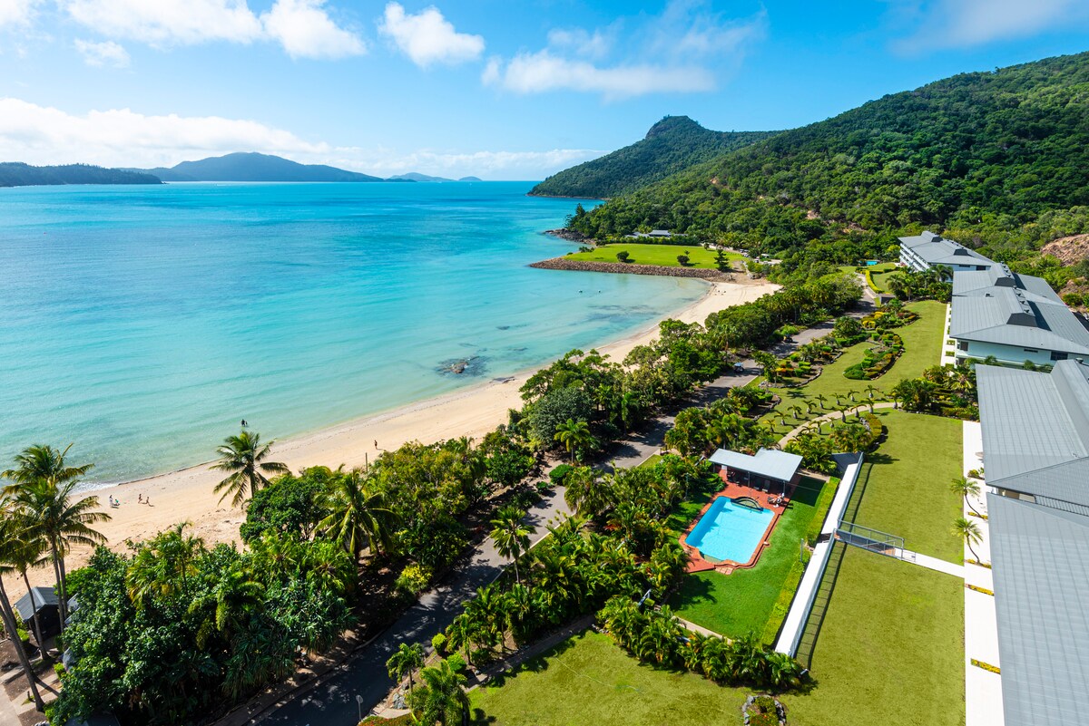 An aerial view captures a lush coastal landscape, featuring a sandy beach bordered by gentle waves. A turquoise pool is nestled among the greenery, surrounded by palm trees and manicured lawns. The backdrop includes a mountainous terrain under a clear blue sky.
