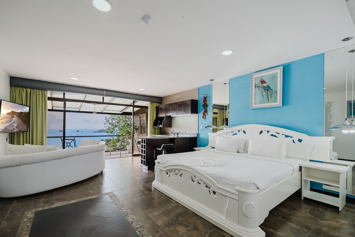 A spacious room features a king-size bed with a decorative headboard and a cozy seating area with a white sofa. Large glass doors open to a terrace, enhancing the unobstructed ocean view. A well-equipped kitchen area is visible, complemented by blue and green accents on the walls.