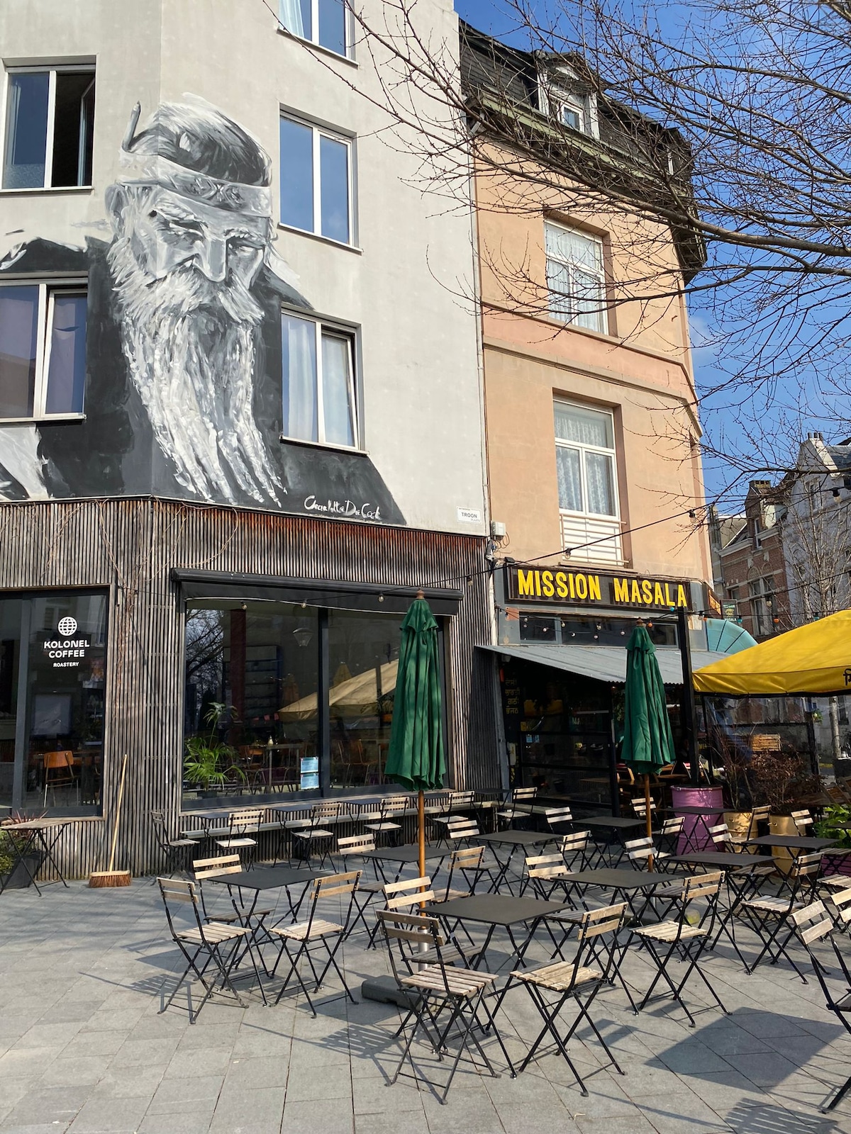 An outdoor seating area is visible, featuring several wooden tables and folding chairs arranged on a cobbled square. A restaurant with a vibrant yellow awning displays its name, while a large mural of an elderly man adorns the building’s facade above.