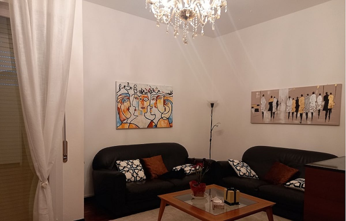 A well-lit living area features two dark leather sofas adorned with decorative cushions. A coffee table sits centrally, complemented by a simple flower arrangement. The walls are decorated with artwork, and a stylish chandelier hangs overhead, providing a warm ambiance.
