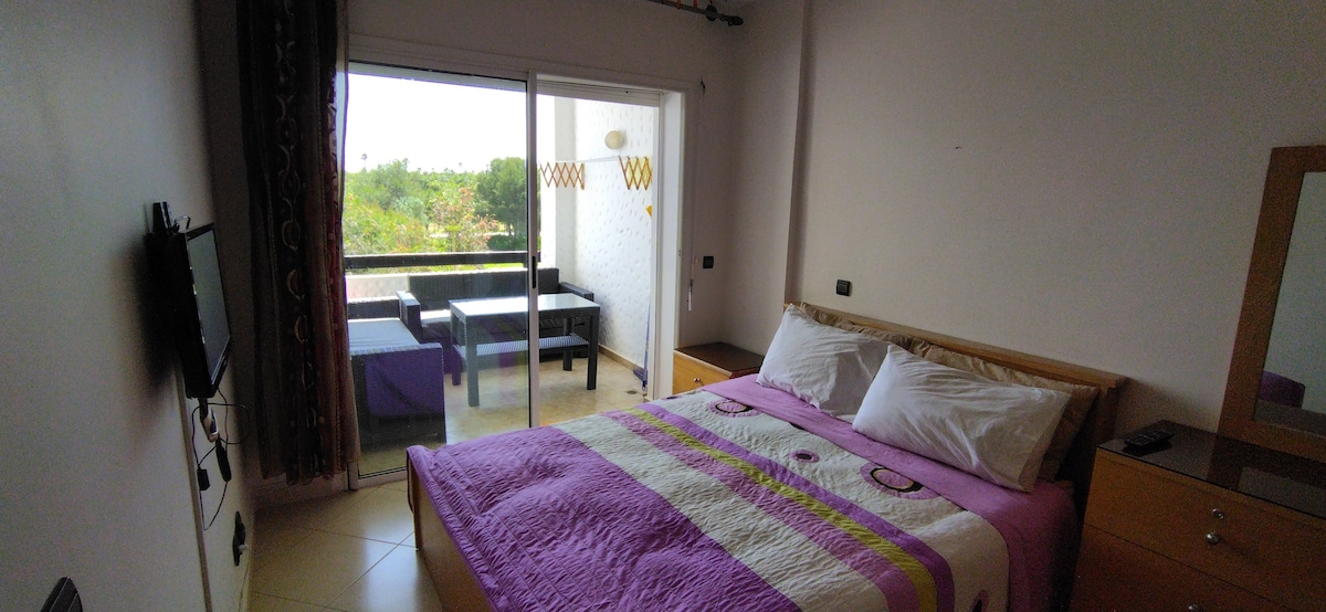 A well-lit bedroom features a spacious bed with a purple quilt, complemented by a matching decorative element. Floor-to-ceiling sliding doors lead to a private balcony area furnished with seating, allowing for a view of the outdoors. A flat-screen TV is mounted on the wall.