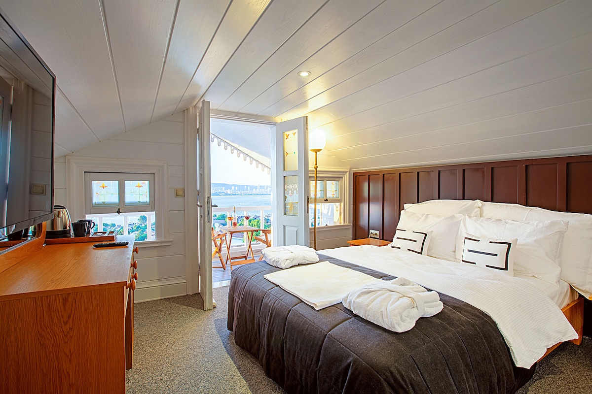 A spacious bedroom is presented, featuring a large bed with crisp white linens. Natural light filters through large windows, with a cozy seating area visible outside on the terrace. A wooden dresser and complimentary amenities are also included, contributing to a comfortable atmosphere.