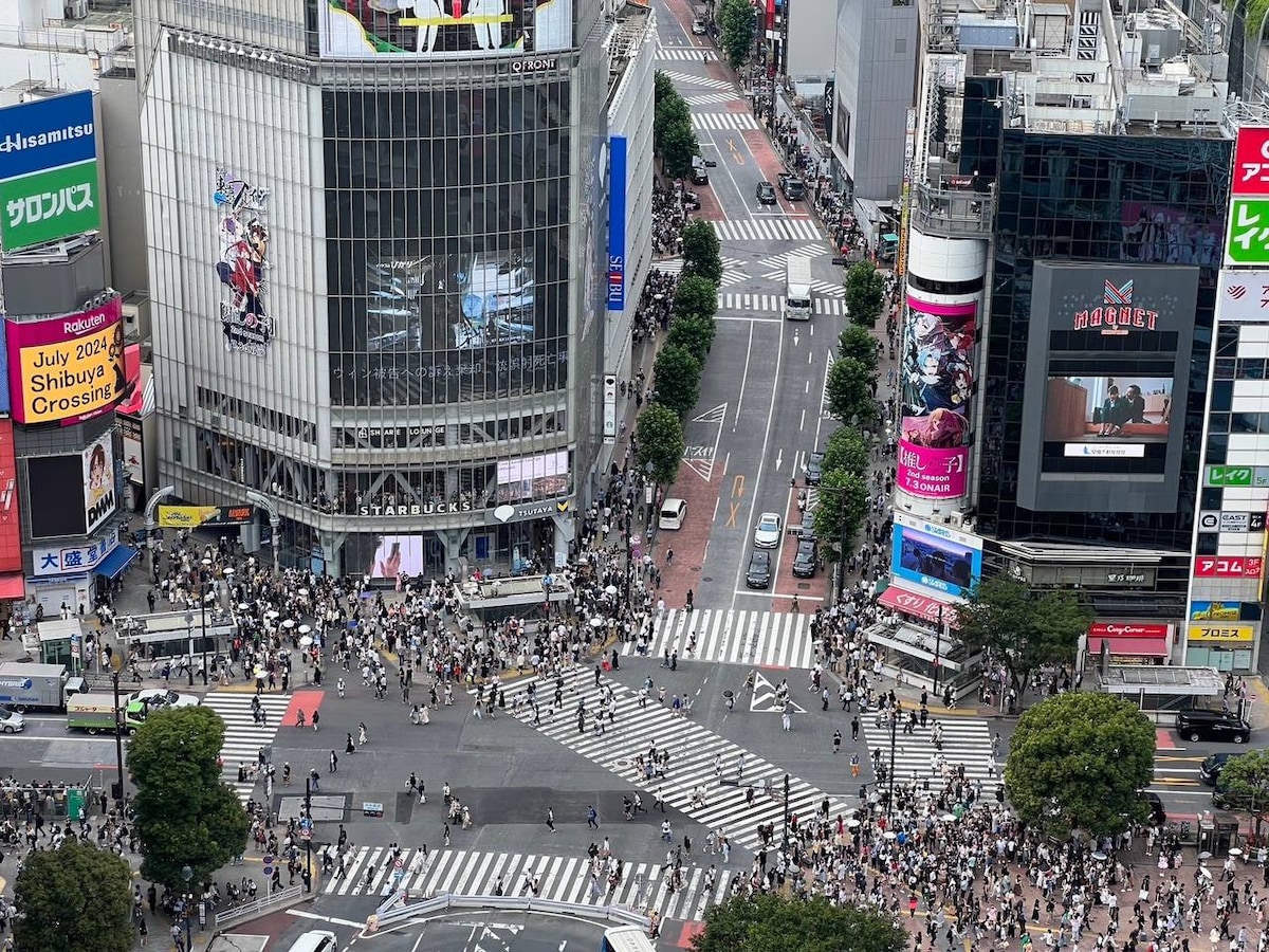 An aerial view captures a bustling urban intersection filled with pedestrians crossing vibrant crosswalks. Surrounding tall buildings feature prominent advertisements and storefronts. The scene reflects the lively atmosphere of the city, with trees and traffic signals adding to the urban landscape.