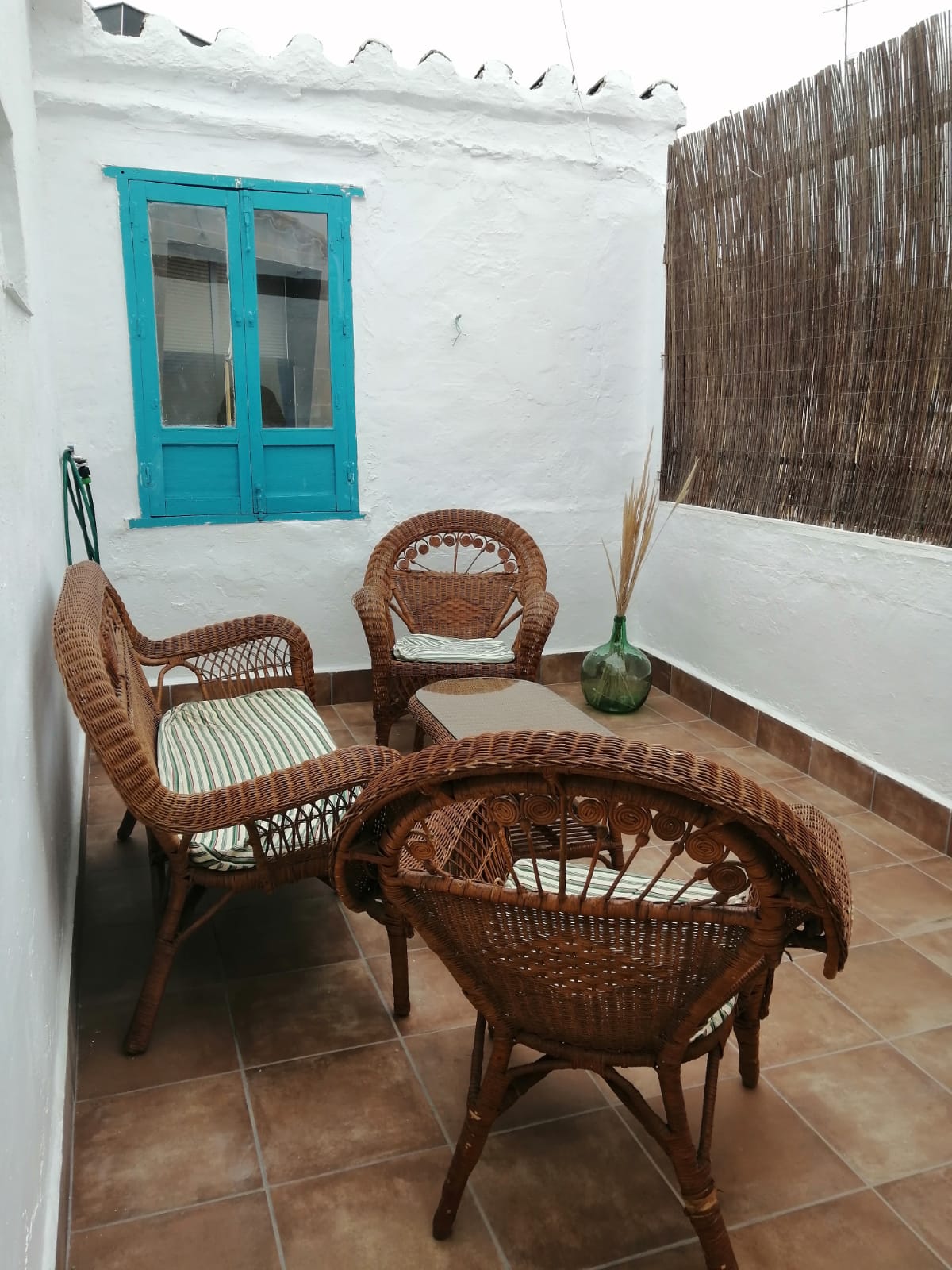 A cozy outdoor seating area is presented with four woven chairs arranged around a small table. A glass vase with decorative accents sits on the table, while a backdrop of textured brown fencing and a bright blue window enhance the inviting atmosphere.