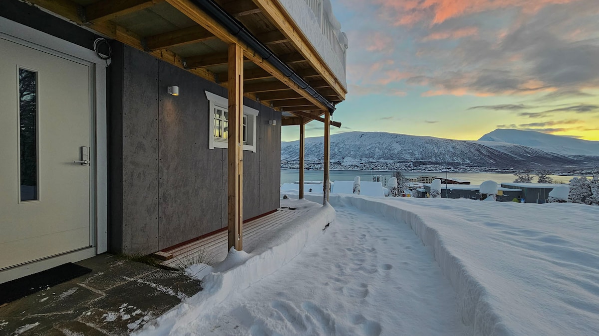 An entrance features a covered porch with wooden beams, leading to a modern door. Snow covers the ground and partially envelops the area, with distant mountains visible against a colorful sunrise sky.