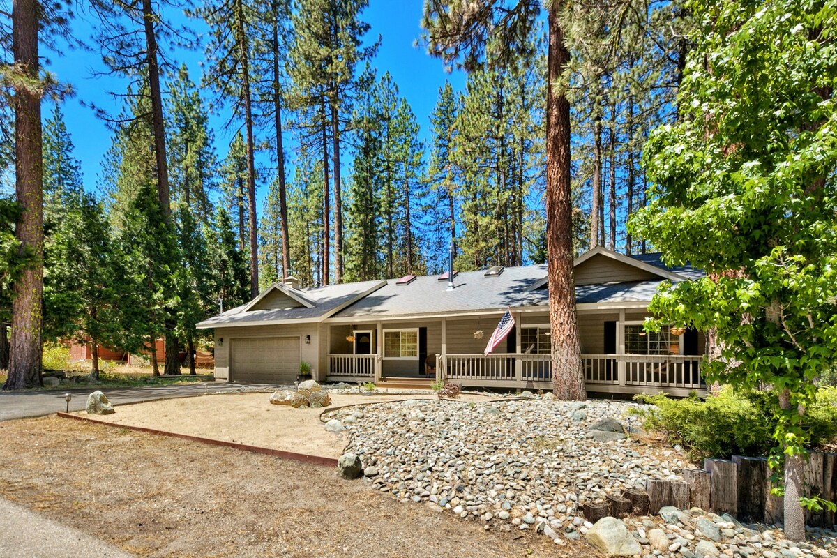 A charming single-story home is framed by tall pine trees, showcasing a welcoming front porch with railings. The exterior features a gravel driveway and a landscaped area with decorative stones and shrubs. An American flag is displayed, enhancing the inviting curb appeal.